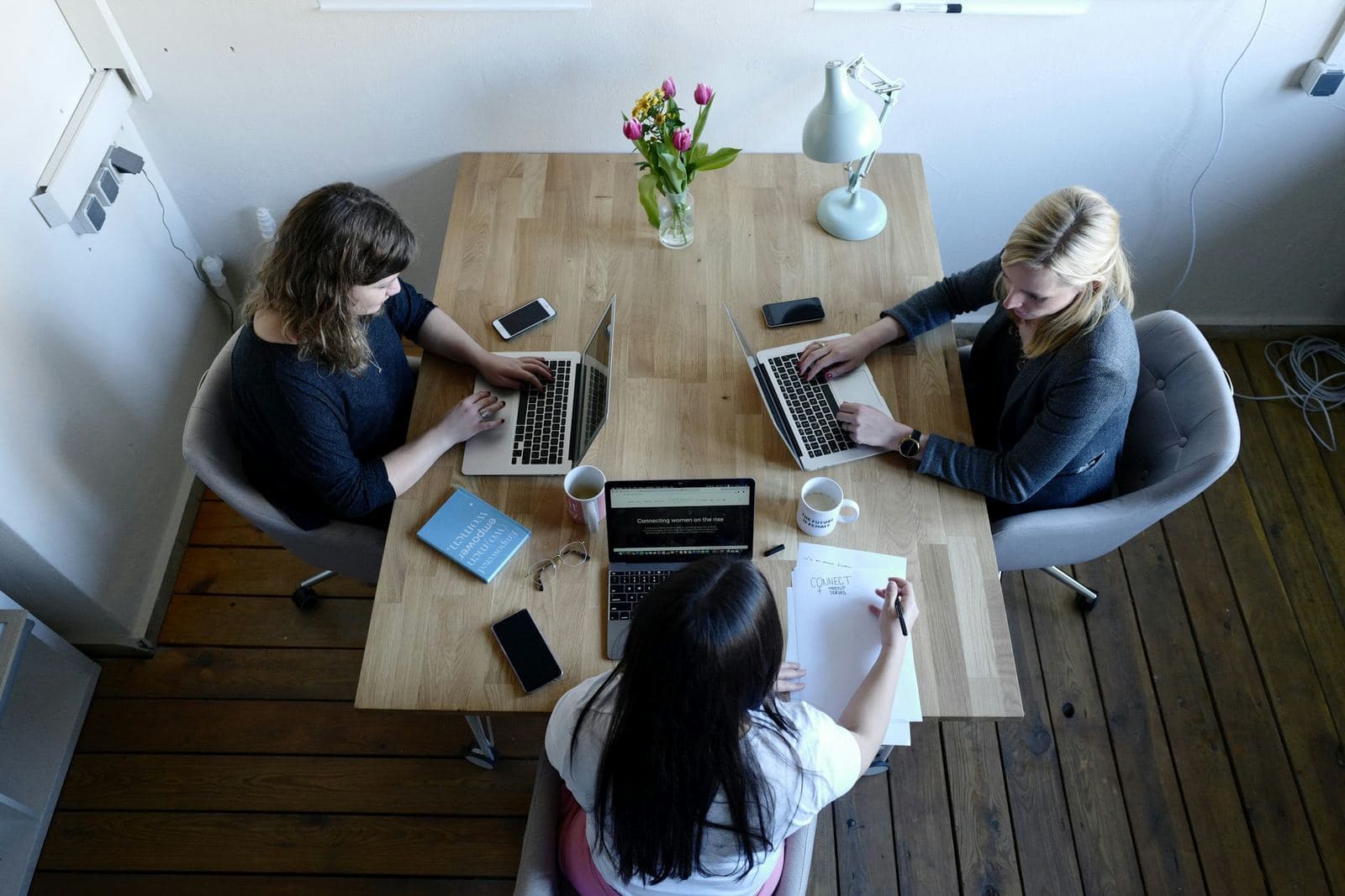 Drie vrouwen werken samen aan een houten tafel met laptops en notities, van bovenaf gezien